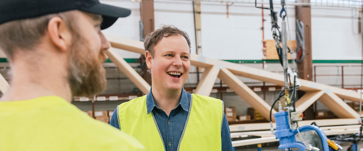 Två män i varselkläder ståendes inne i en produktion där man kan skymta takstolar i bakgrunden. 