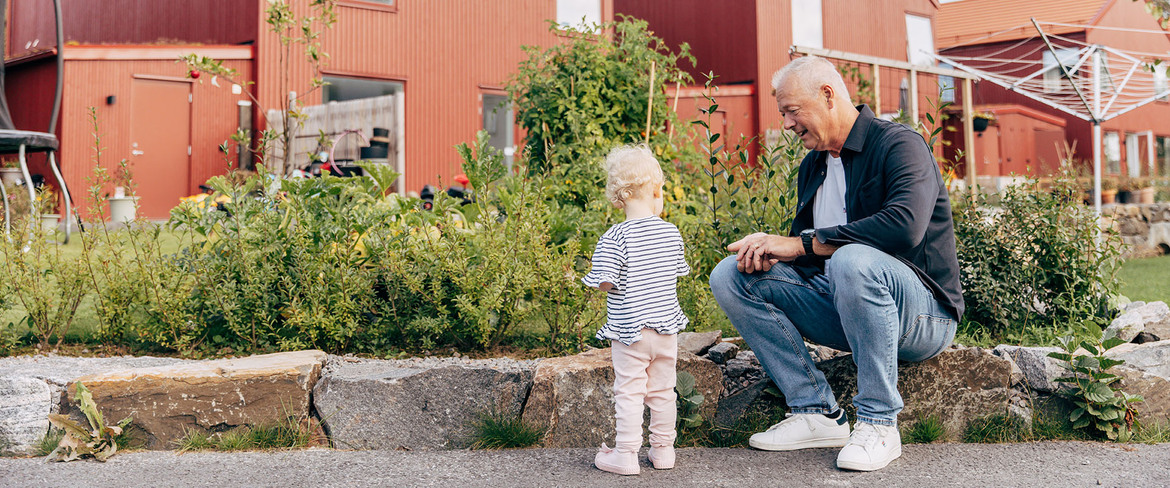 En man sitter på en stenmur och samtalar med ett barn framför ett rött villaområde byggt i trä. 