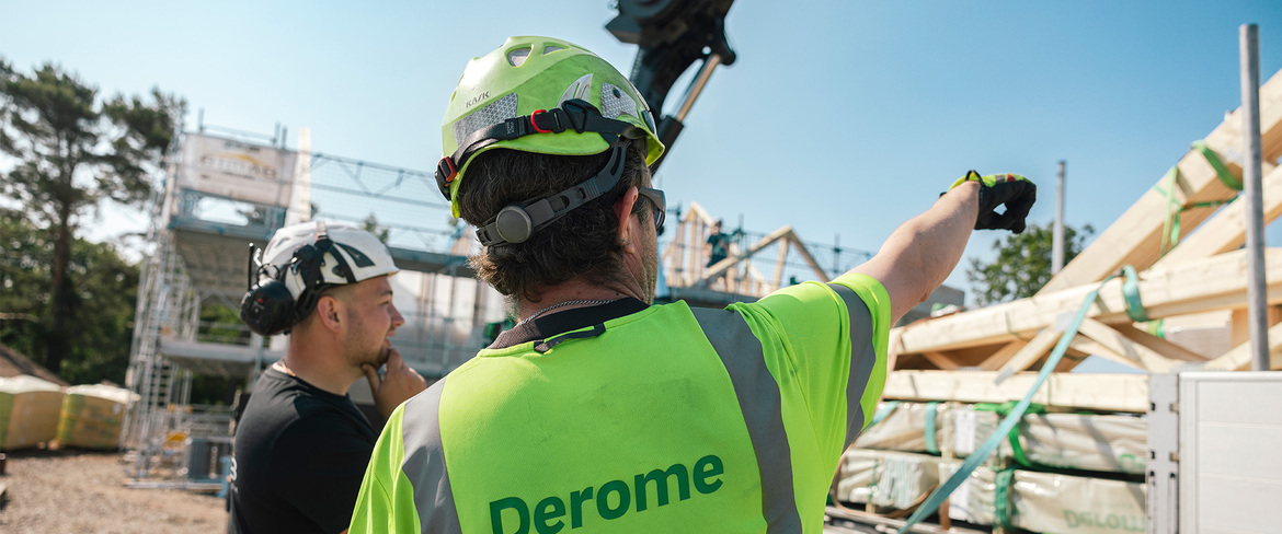 En man i varselkläder pekar mot träkonstruktioner som ligger fastbundna i en kranbil som ska lastas av vid en byggnadsplats. 